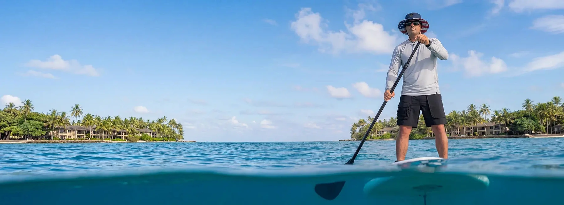 Paddleboarder wearing outdoor apparel and a boonie hat on clear blue water