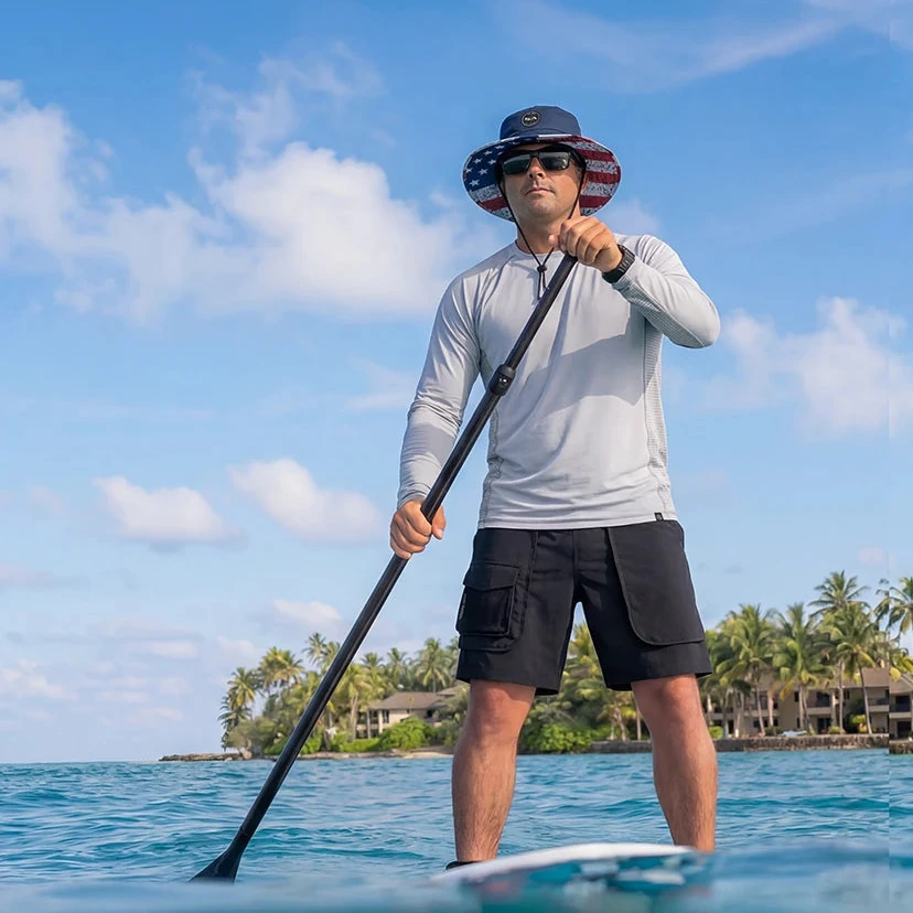 Paddleboarder wearing outdoor apparel and a boonie hat on clear blue water
