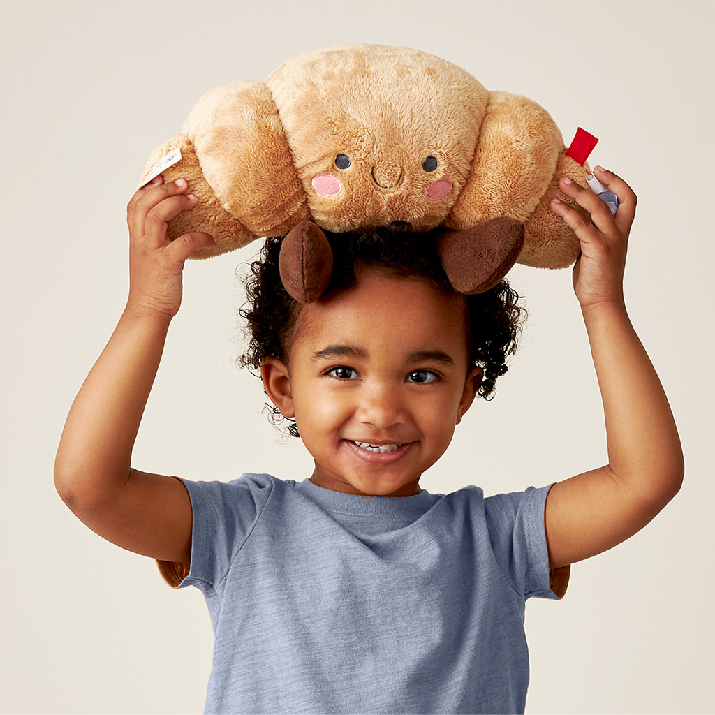Child holding an Itzy Ritzy plush croissant toy overhead